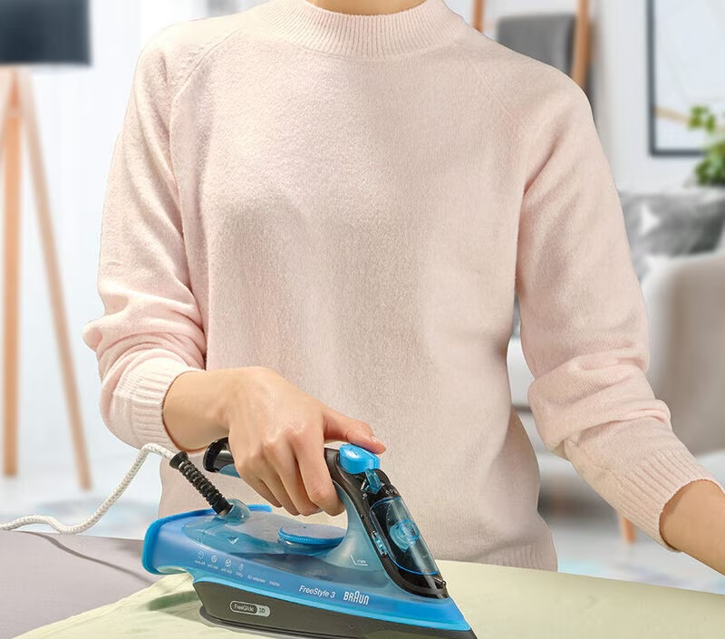 Osoba prasuje ubrania niebiesko-czarnym żelazkiem. Ma na sobie różowy sweter. Tło przedstawia pokój z meblami.
