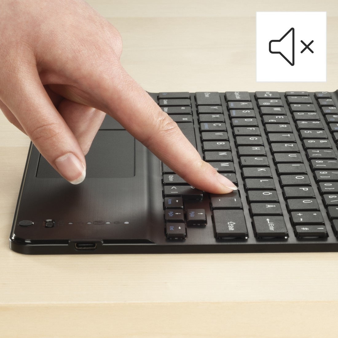 Ein Finger drückt eine Taste auf einer schwarzen Tastatur mit Touchpad. Ein Ton-aus-Symbol ist sichtbar.