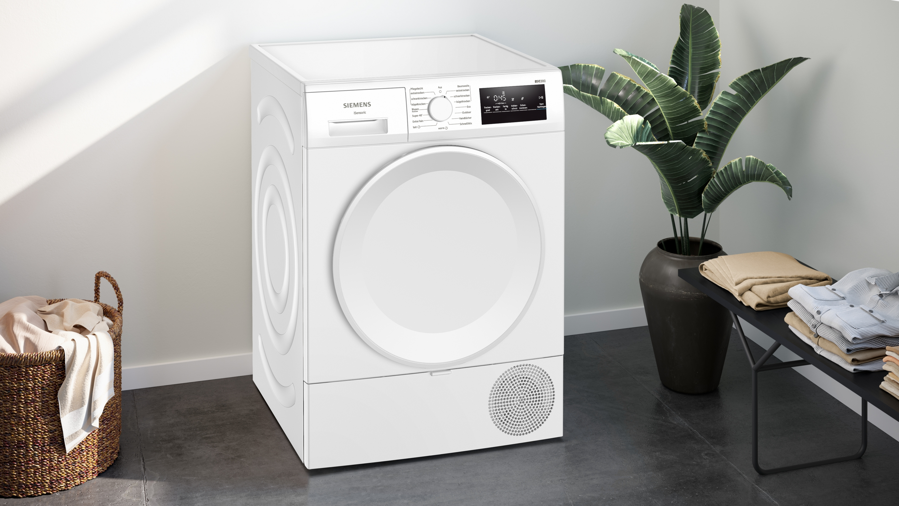 Ein weißer Siemens-Trockner steht in einem Raum neben einer Pflanze in einem Topf, einer Bank mit Kleidung und einem Wäschekorb.