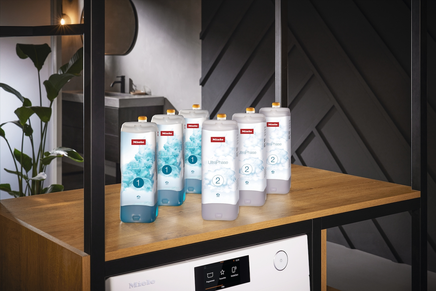 Mehrere Miele UltraPhase Waschmittelflaschen stehen auf einem Holzregal. Darunter eine Waschmaschine. Der Hintergrund zeigt ein Badezimmer.