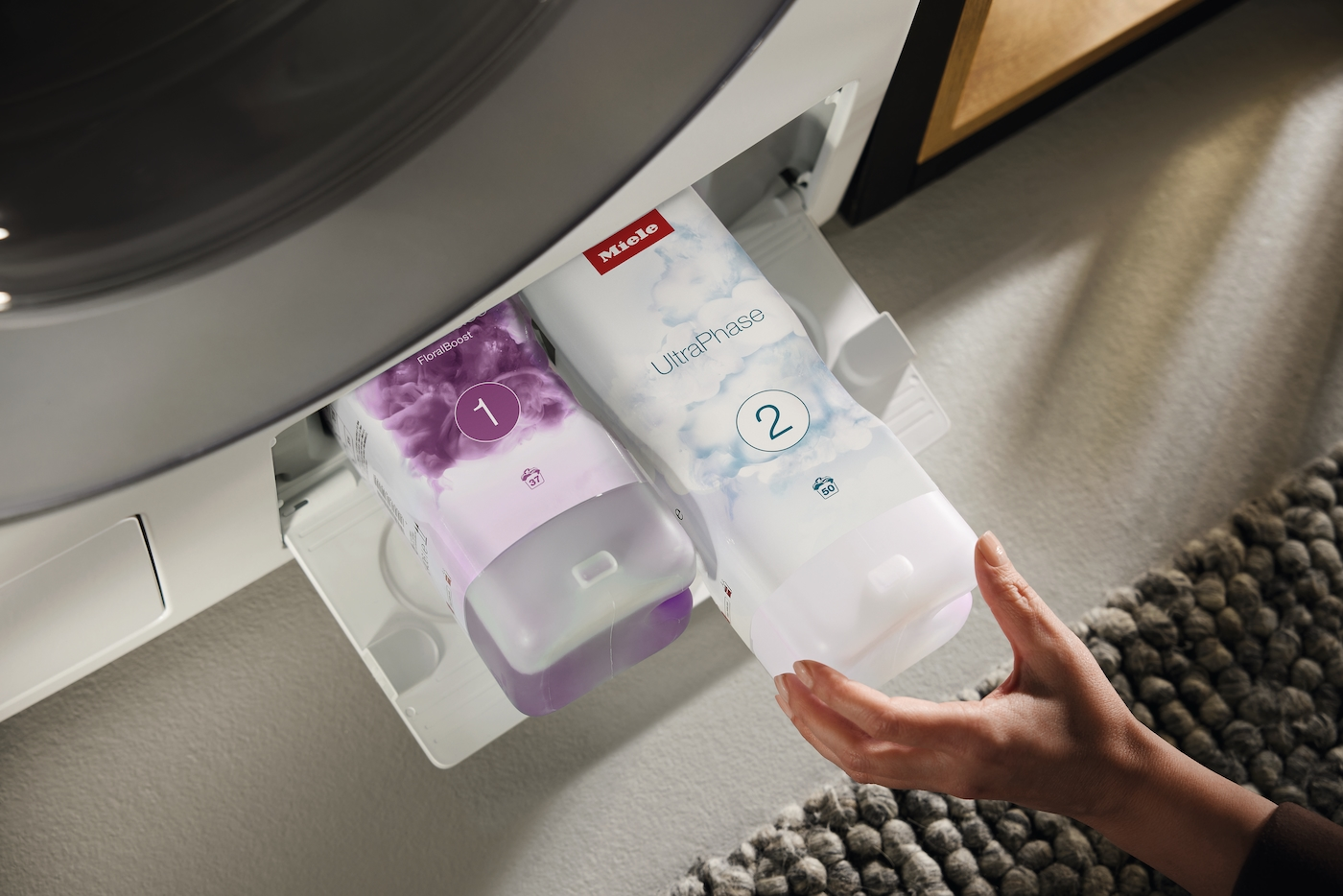 Eine Hand hält einen Waschmittelbehälter. Eine Waschmaschinenlade enthält zwei Waschmittelkartuschen mit der Aufschrift 'FloralBoost' und 'UltraPhase'.
