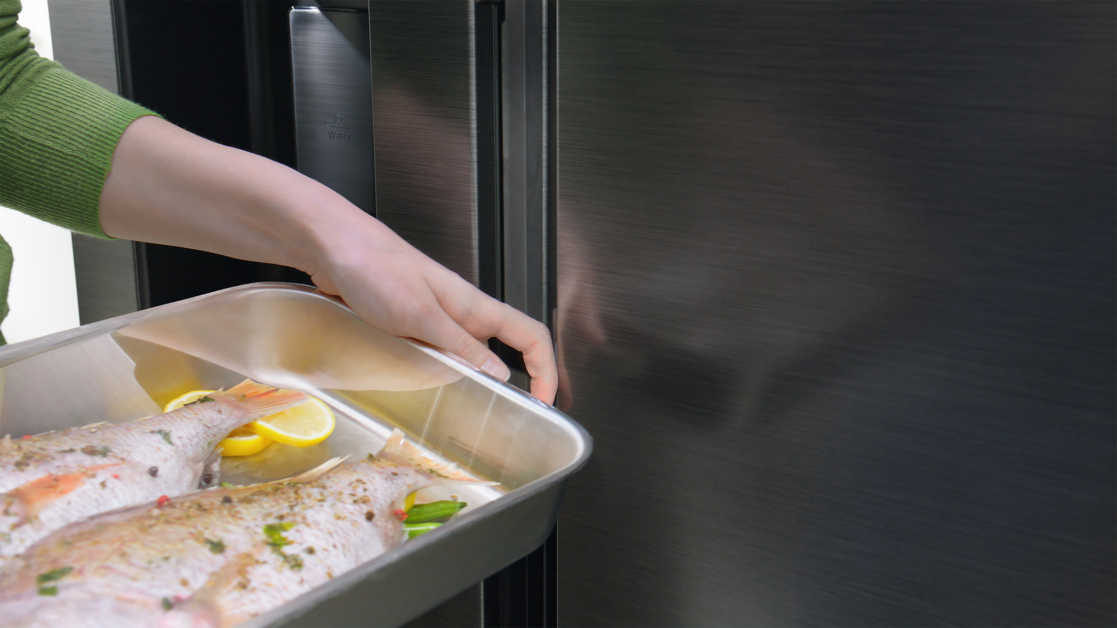 Hand hält ein Tablett mit Fisch und Zitronen neben einer dunklen Kühlschranktür.