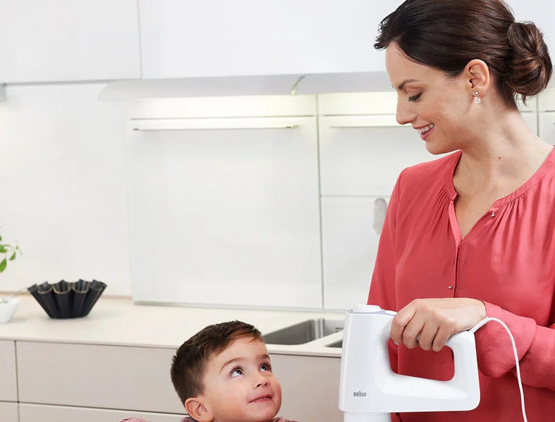 Kobieta trzyma mikser patrząc na dziecko. Tło kuchni z białymi szafkami.