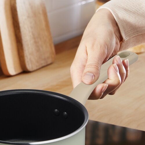 Ręka trzyma uchwyt małego, jasnozielonego rondla. Tłem jest drewniana powierzchnia.