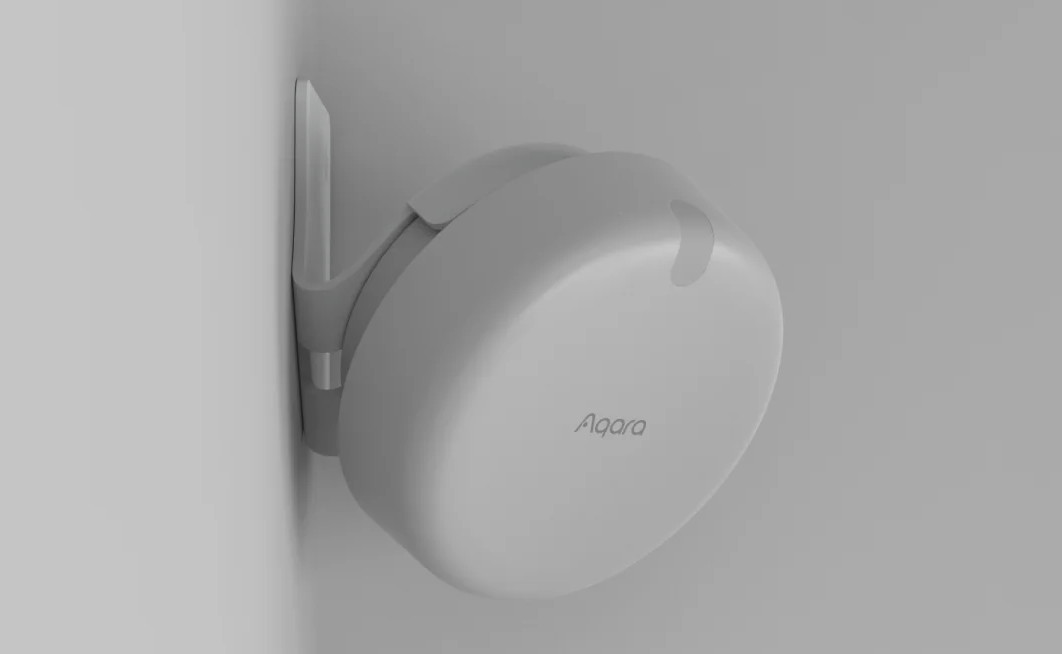 Białe urządzenie smart home Aqara, zamontowane na białej ścianie. Urządzenie jest okrągłe i ma logo Aqara.