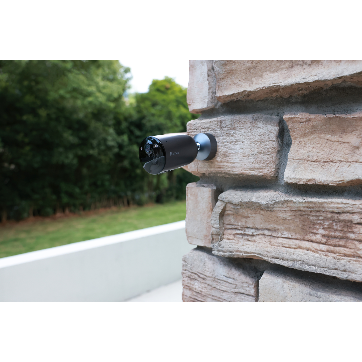 A black security camera mounted on a brick wall, outdoors in a green environment.