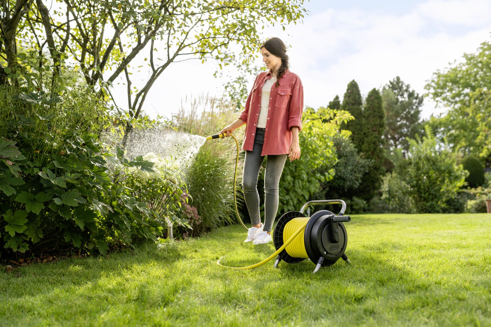 Frau bewässert Pflanzen mit einem Schlauch in einem grünen Garten. Schlauchtrommel liegt auf dem Rasen. Bäume und Büsche im Hintergrund.
