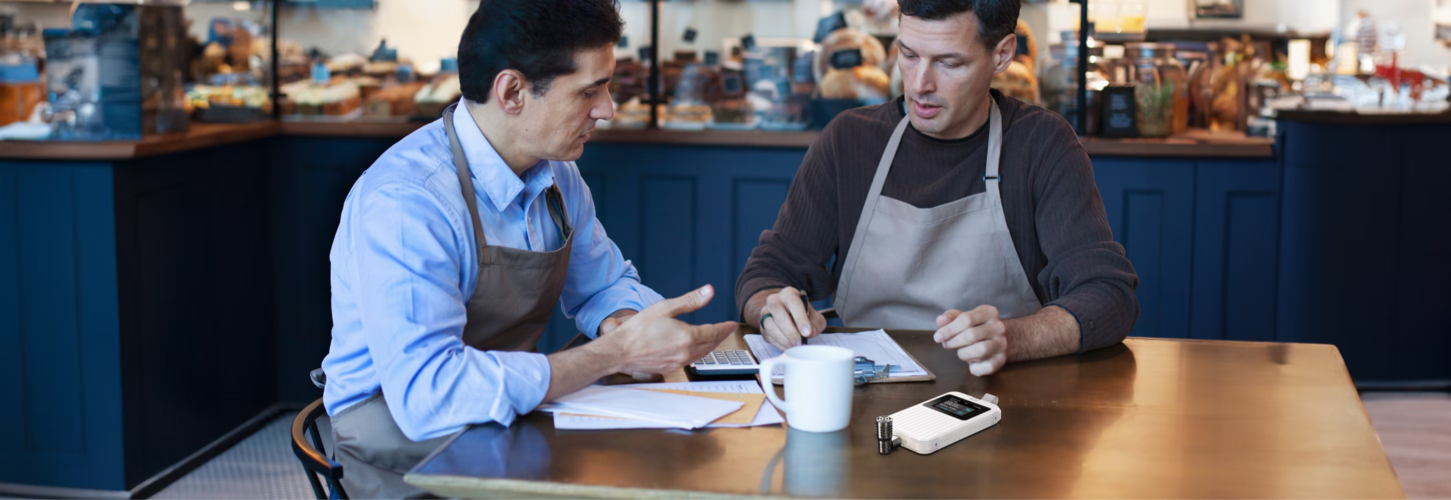 Deux personnes dans un café discutant de documents. Sur la table en bois, une tasse de café, un stylo et un petit appareil.