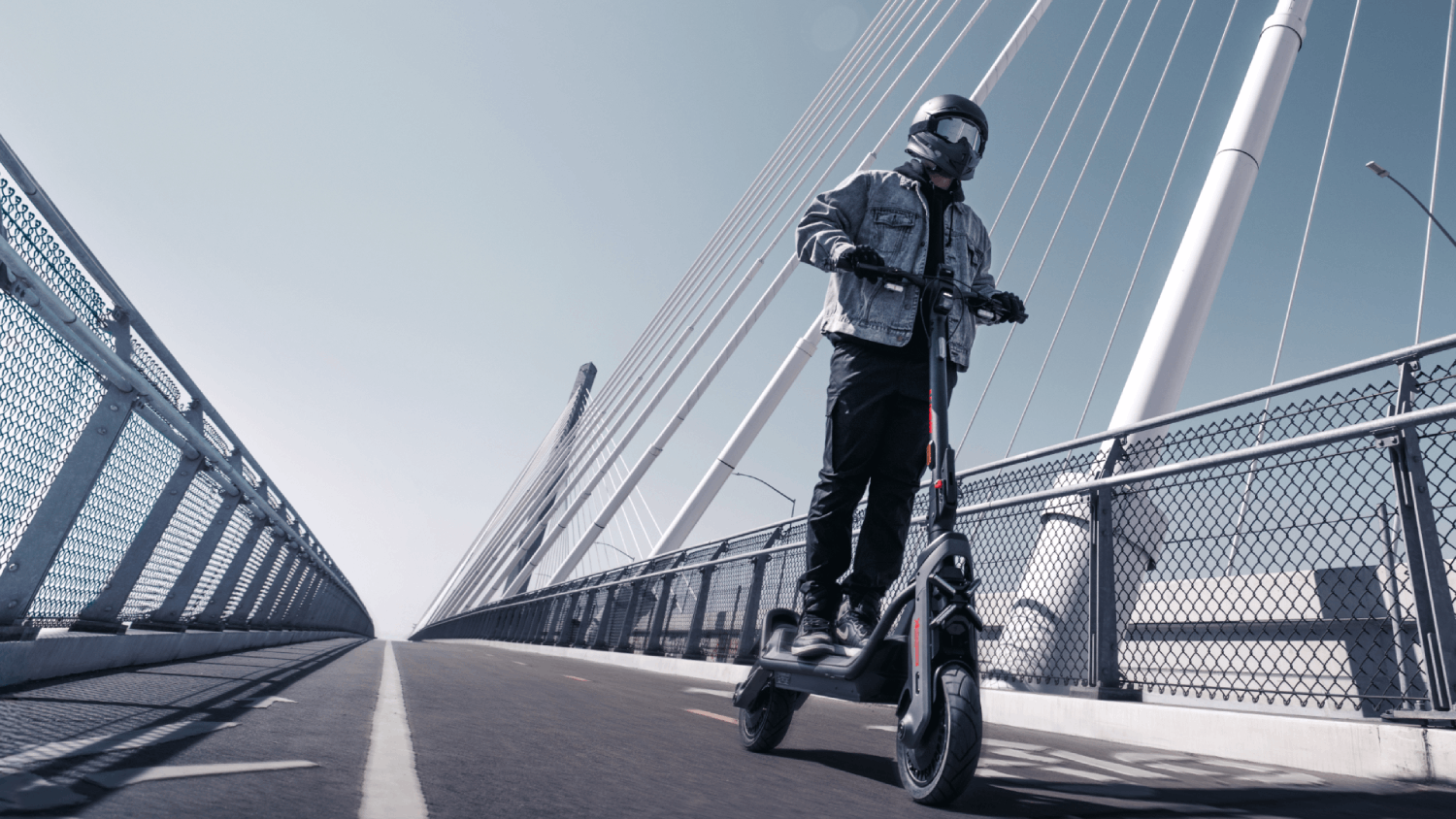 Person auf einem Roller fährt über eine Brücke. Die Person trägt einen Helm und eine Jacke. Der Himmel ist blau.