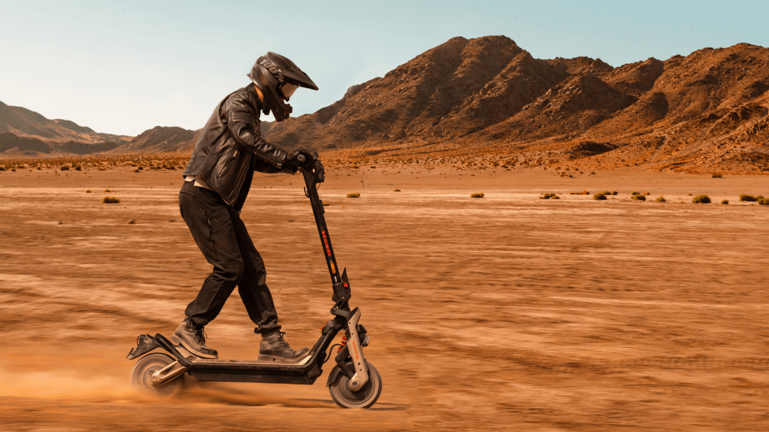 Persona de negro montando un patinete en el desierto. Montañas al fondo y cielo azul.