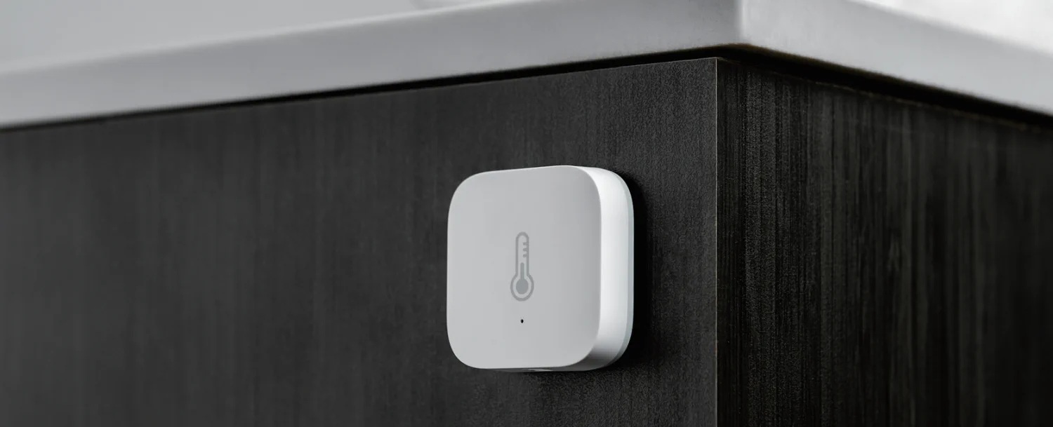 Biały czujnik smart home zamontowany na ciemnej drewnianej powierzchni. Posiada ikonę termometru.