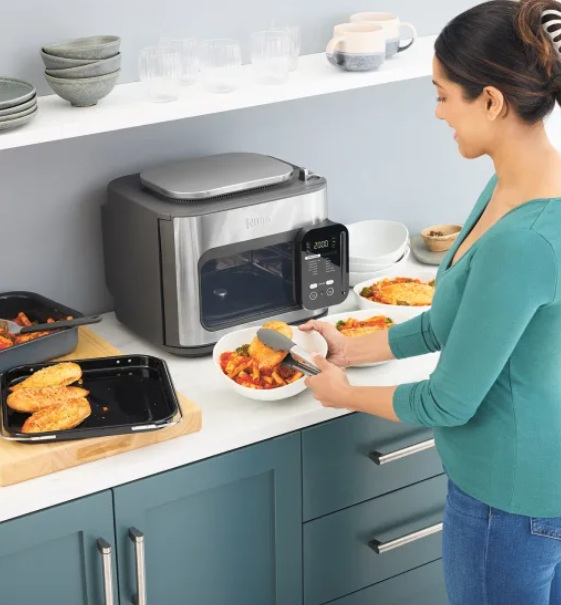 Kobieta umieszcza jedzenie z tacy do miski. Piekarnik i inne naczynia na blacie.