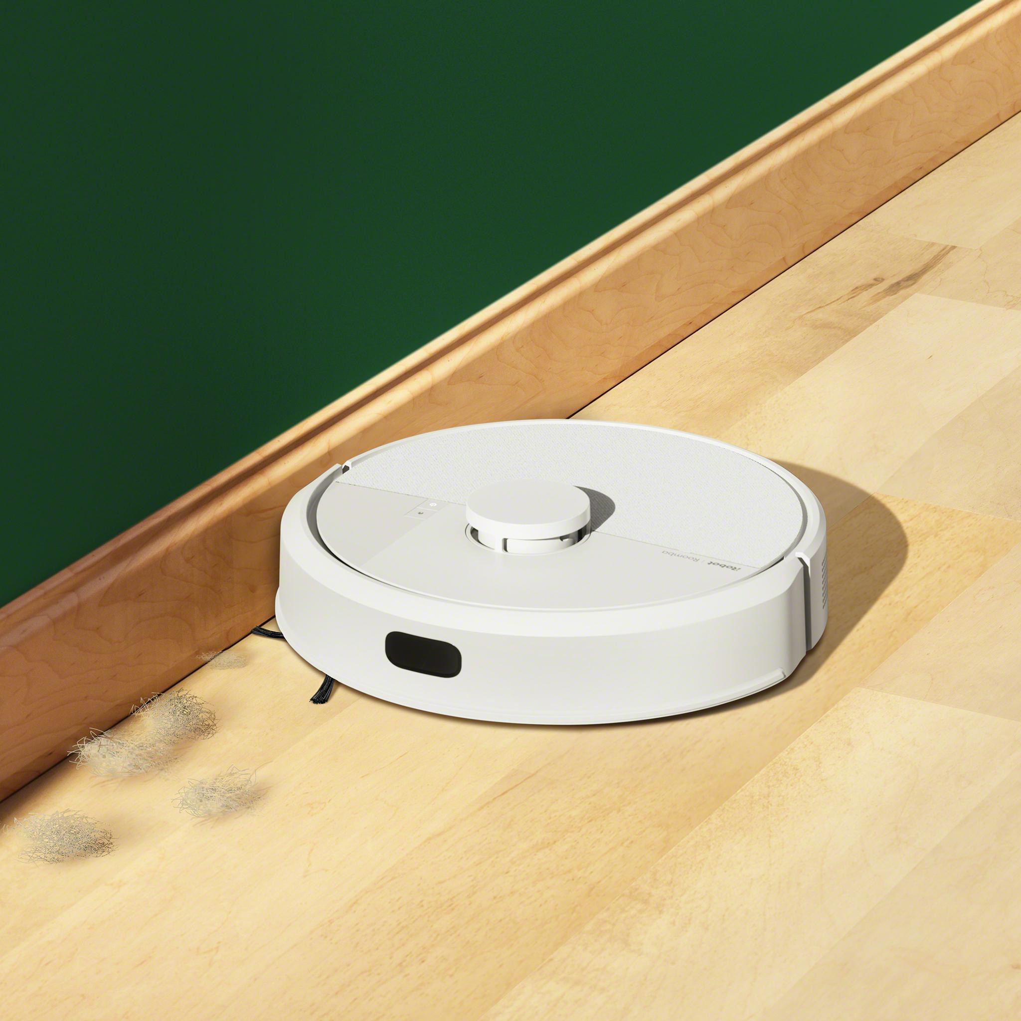 Ein weißer Roboter-Staubsauger auf einem Holzboden, neben einer grünen Wand und einem Holzrahmen.
