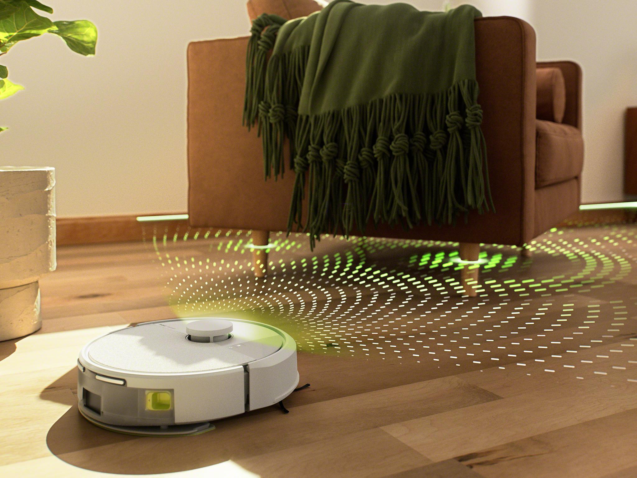 Roboter-Staubsauger auf Holzboden. Im Hintergrund ein brauner Sessel und eine grüne Decke.