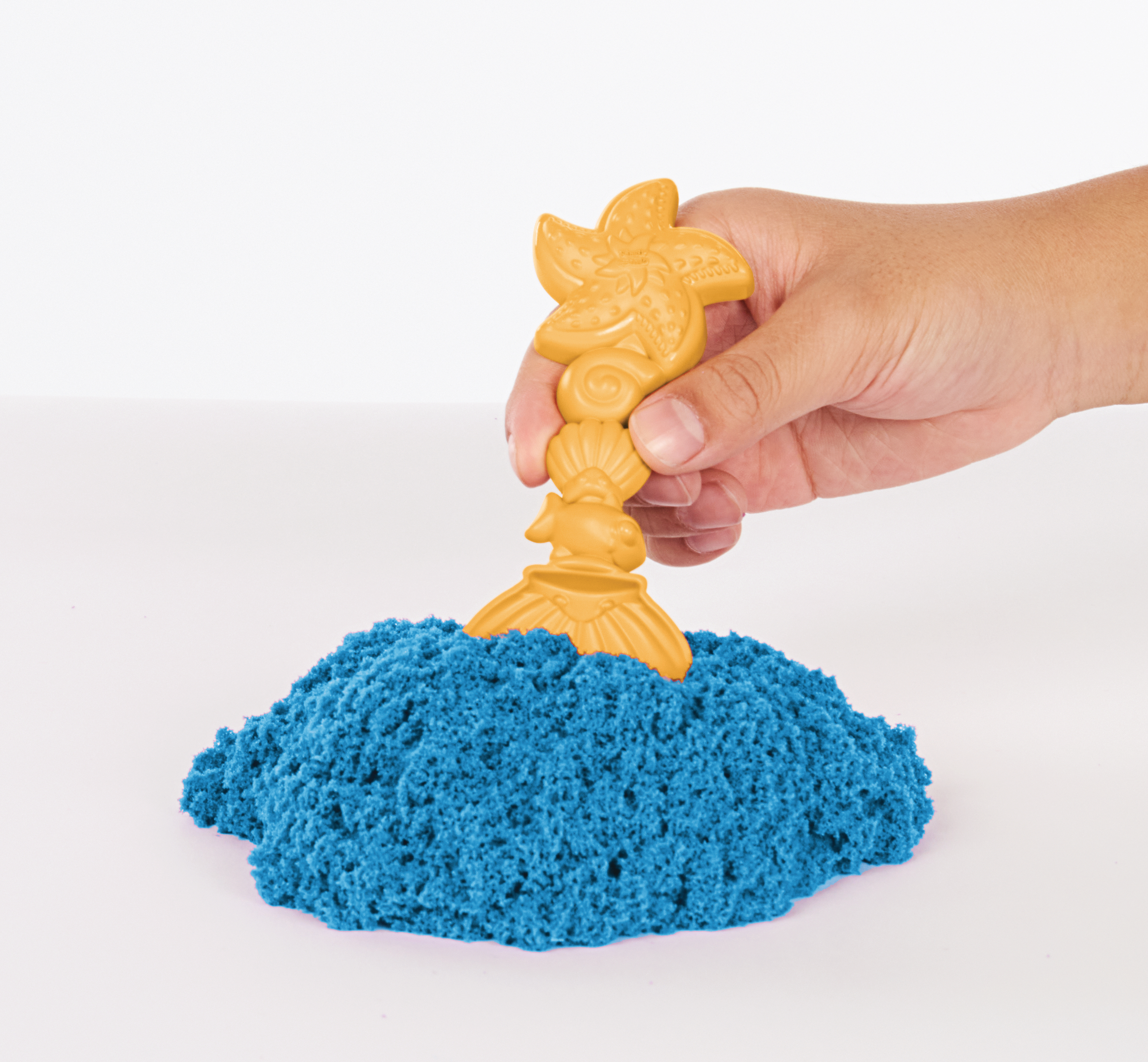 Hand hält gelbes, sternförmiges Sandwerkzeug in blauem Kinetiksand, weißer Hintergrund.