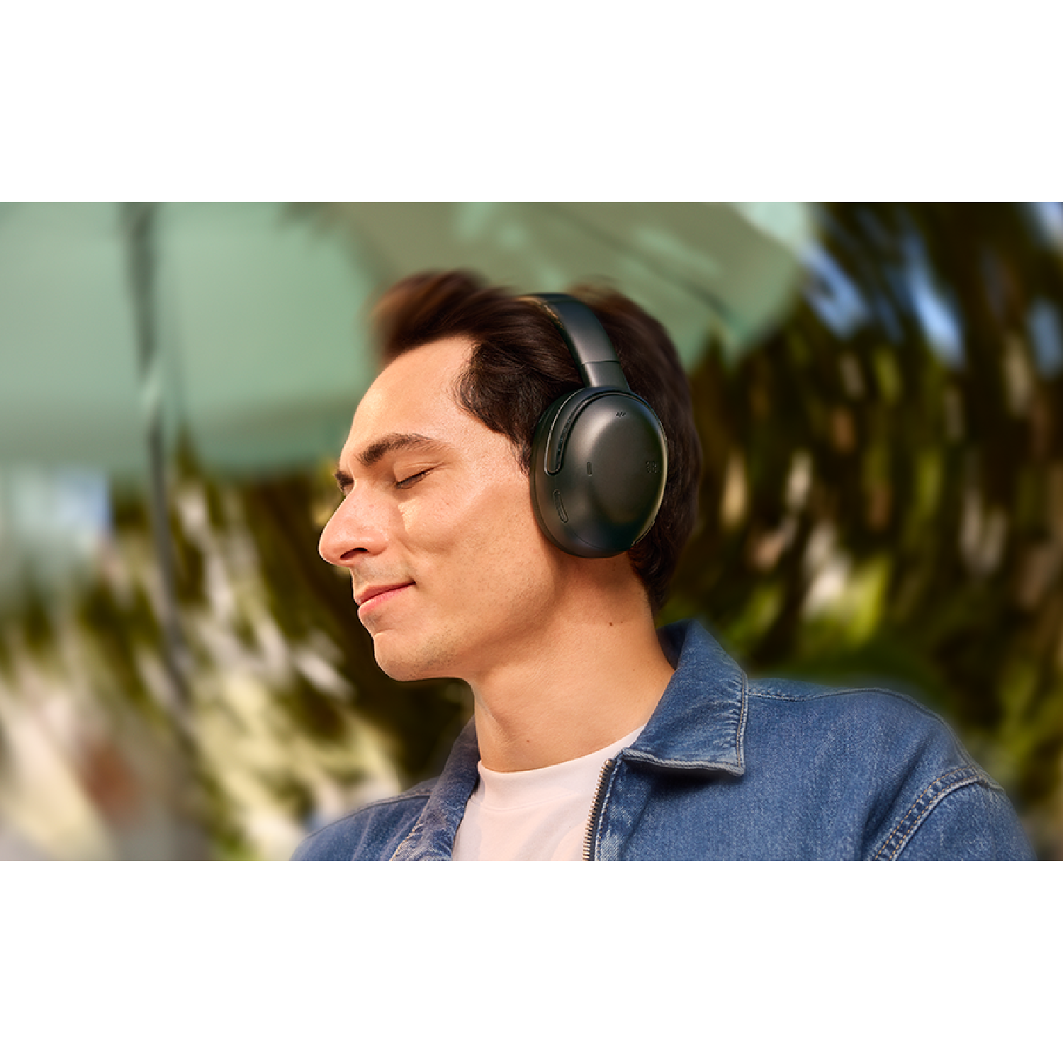 A man with closed eyes wearing black headphones, blurred background, denim jacket.