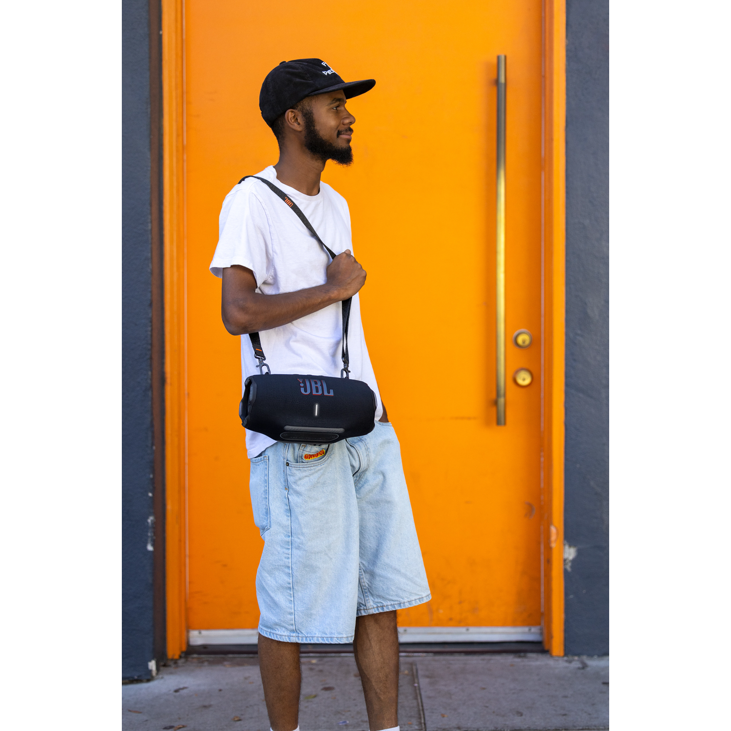 Man wearing a cap and carrying a black JBL speaker on his shoulder.