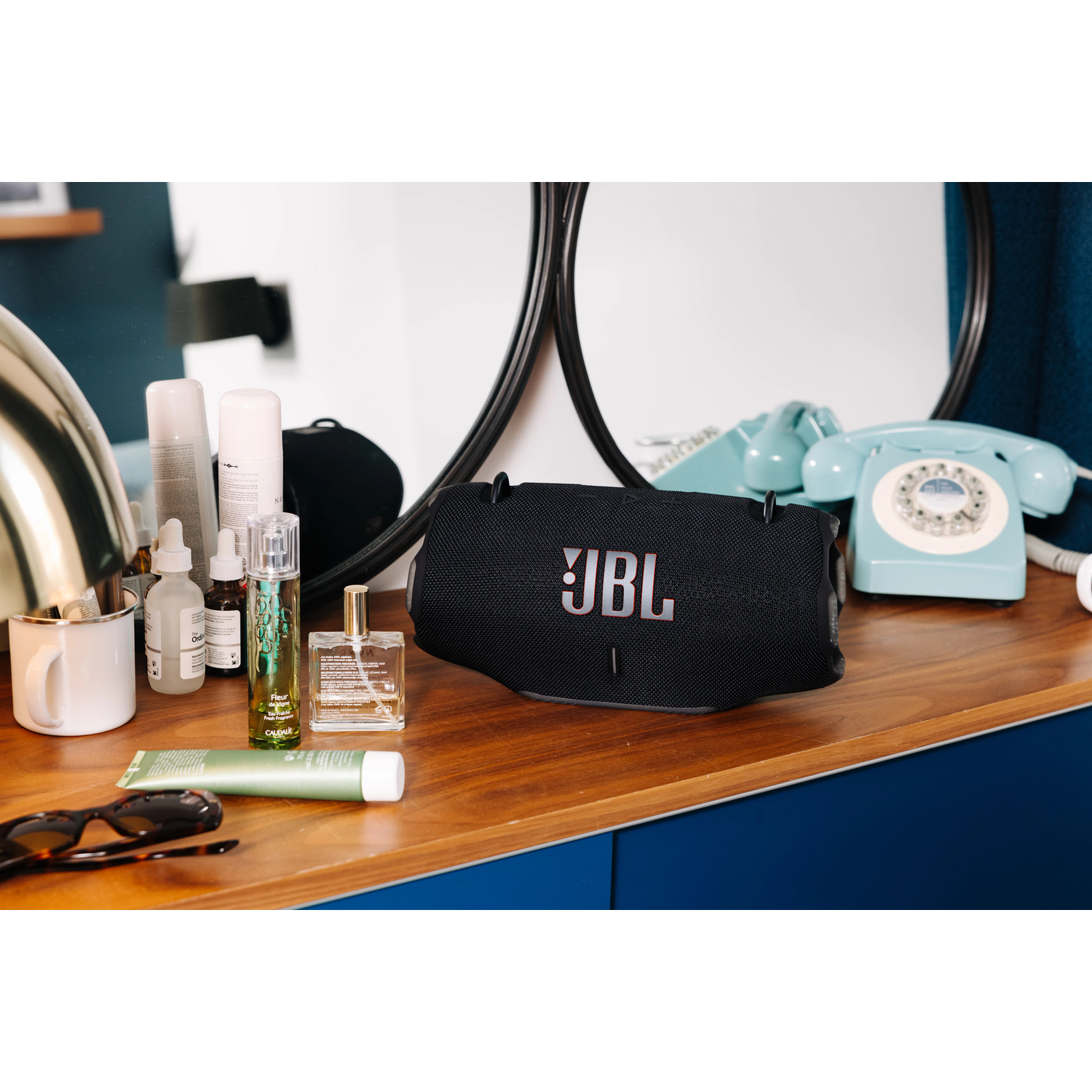 A black JBL speaker sits on a wooden surface with toiletries and a vintage blue telephone. A mirror hangs in the background.