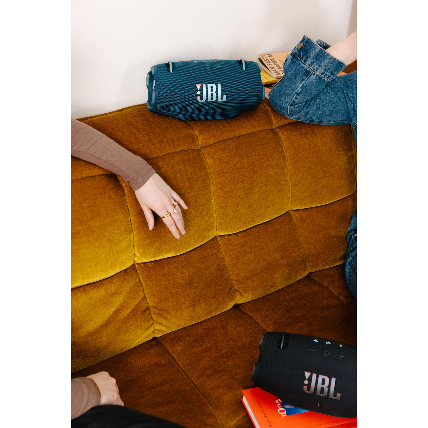 Deux enceintes JBL, une sarcelle, une noire, sur un canapé orange. Mains sur le canapé. Jean en arrière-plan.