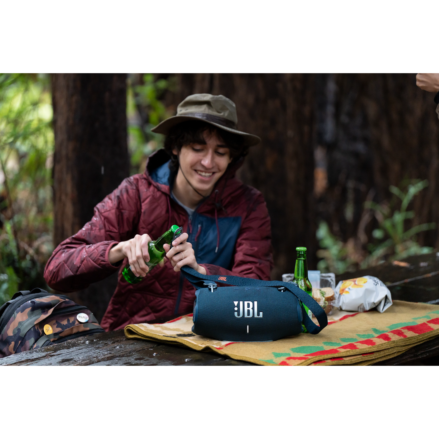 Un homme ouvre une bouteille de bière, avec une enceinte JBL et un sac sur la table. Arrière-plan de forêt.