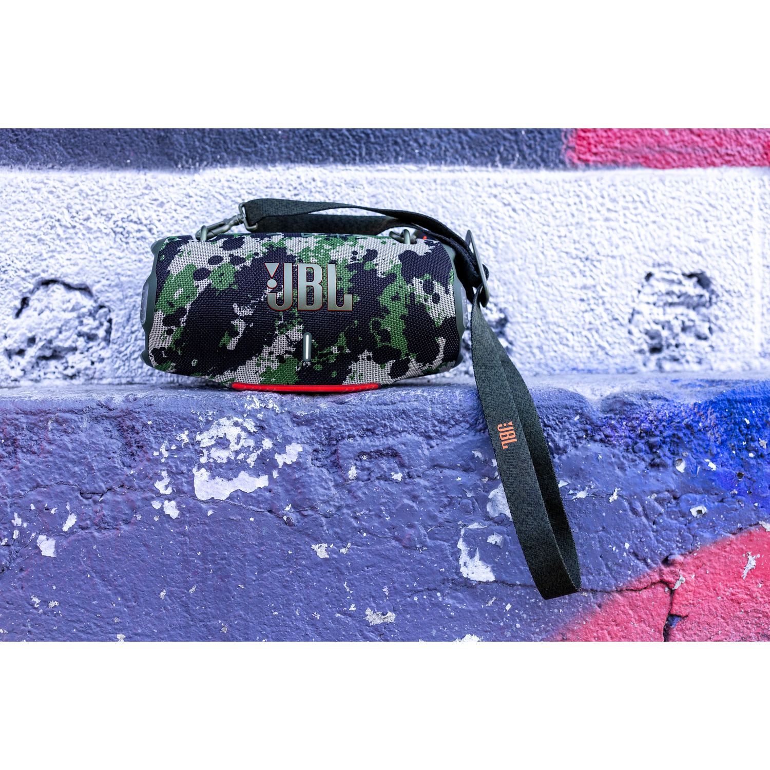 Un haut-parleur JBL avec un motif camouflage repose sur une surface violette contre un mur de graffiti. Une sangle noire pend du haut-parleur.