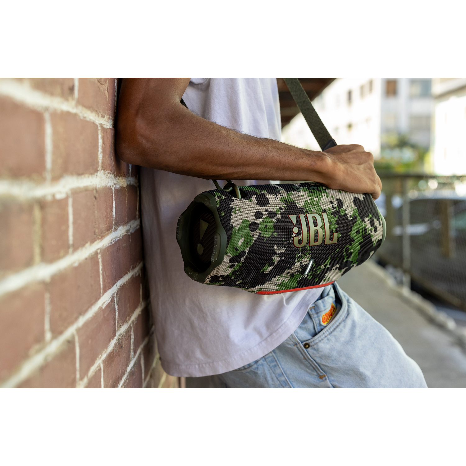 Un homme s'appuie contre un mur de briques, tenant un haut-parleur JBL camouflage. Il porte un t-shirt blanc et un jean. L'arrière-plan montre des bâtiments.