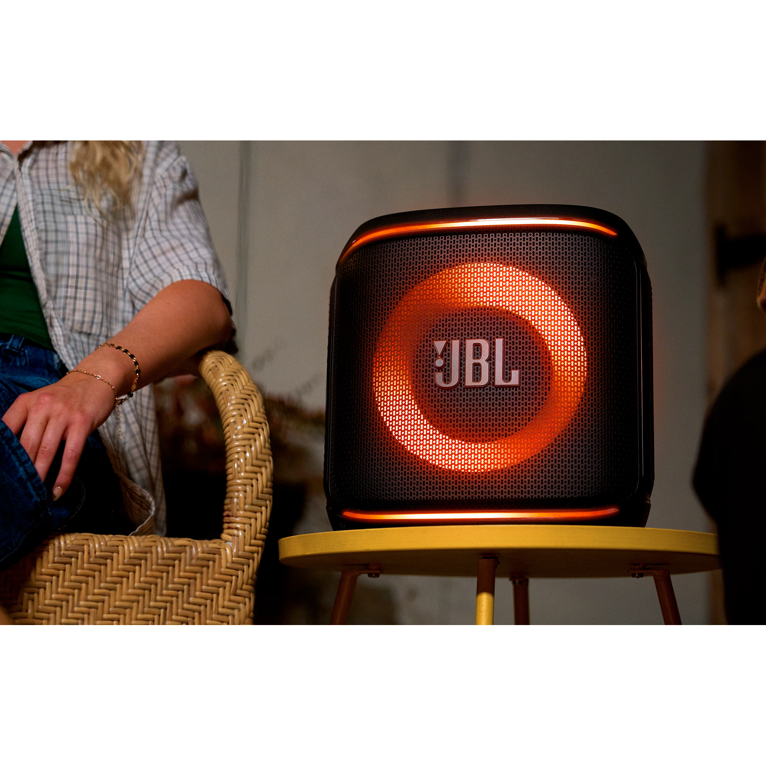 Een JBL-luidspreker met een oranje lichtring staat op een gele tafel. Er staat een persoon in de buurt.