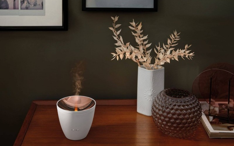 Biały dyfuzor z miedzianą górą, emitujący parę, na drewnianym stole z dekoracjami.