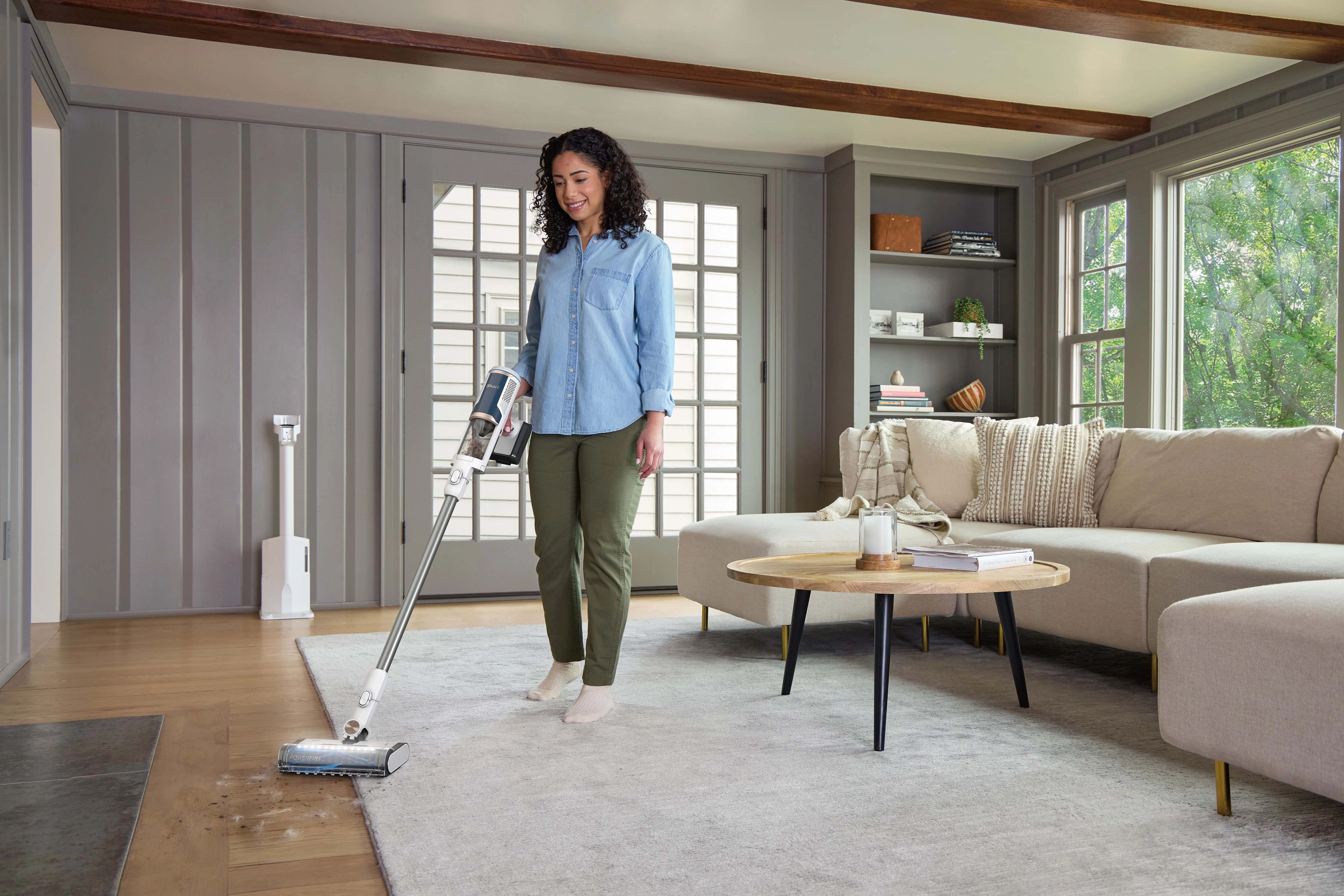 Kobieta odkurza dywan w salonie. Ma na sobie niebieską koszulę i zielone spodnie.
