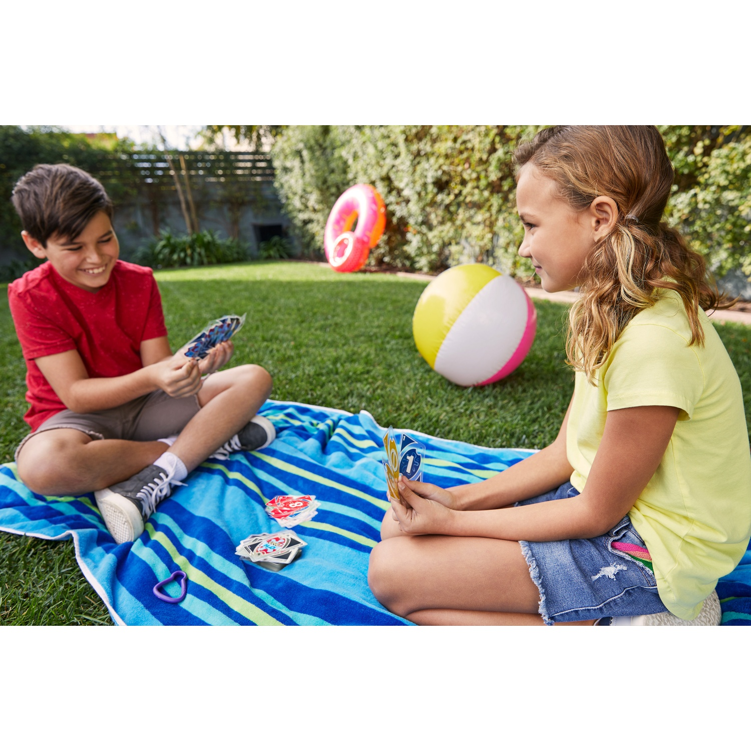 Due bambini che giocano a carte su una coperta. Primo piano: bambini e carte. Sfondo: pallone da spiaggia ed erba verde.