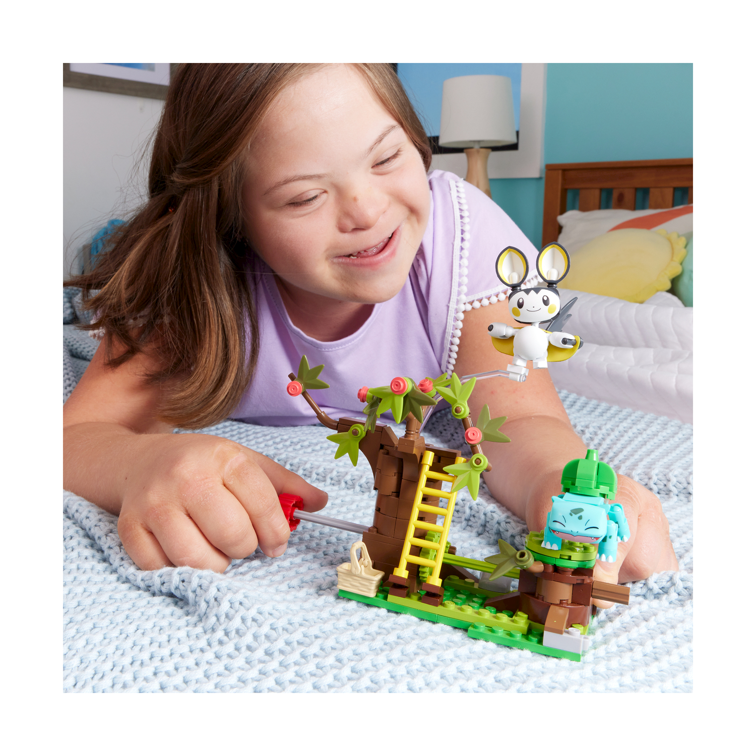 Una bambina con sindrome di Down sorride, costruendo una scena Lego con albero e personaggi.