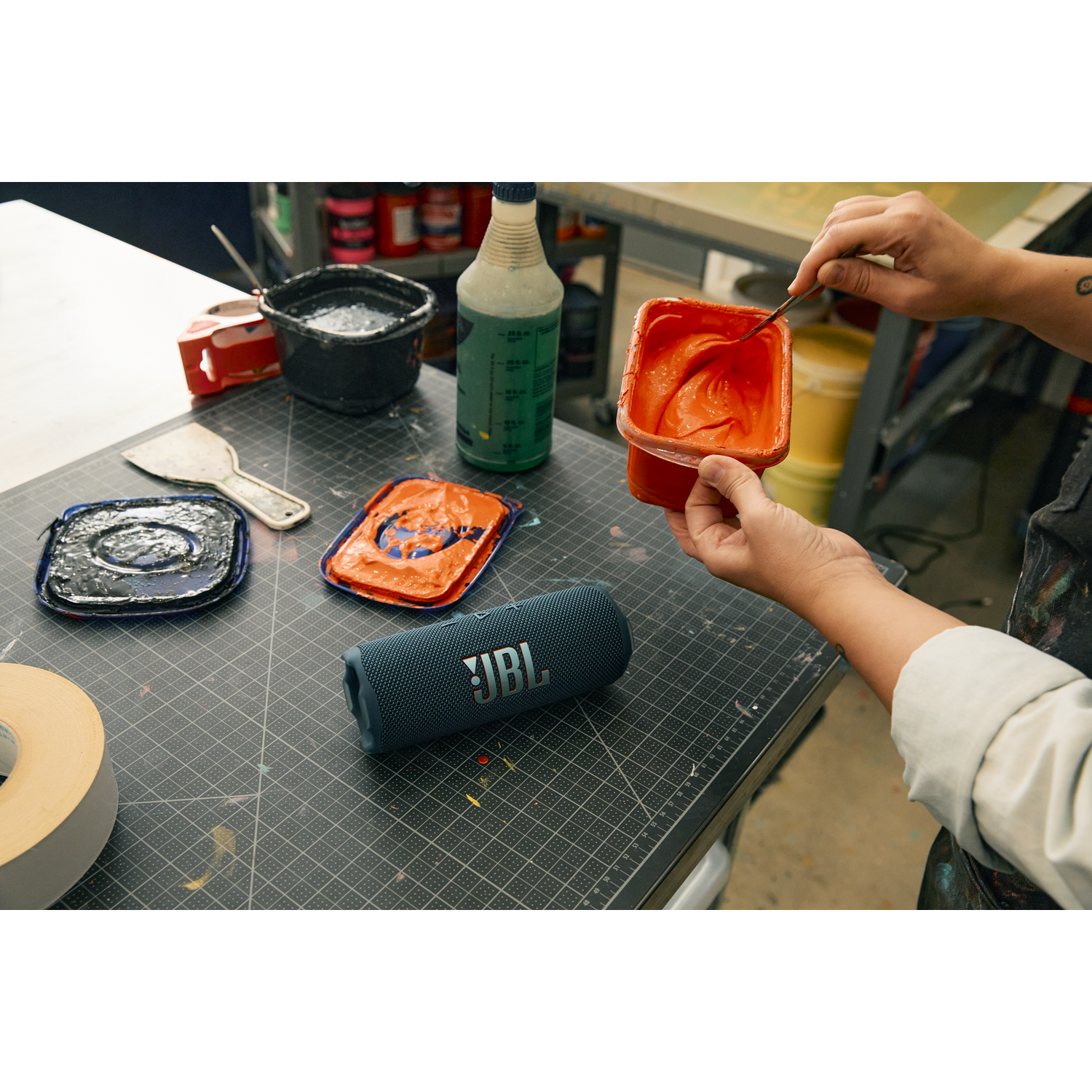 Hands mixing orange paint. A blue JBL speaker and other art supplies are on a grey table.
