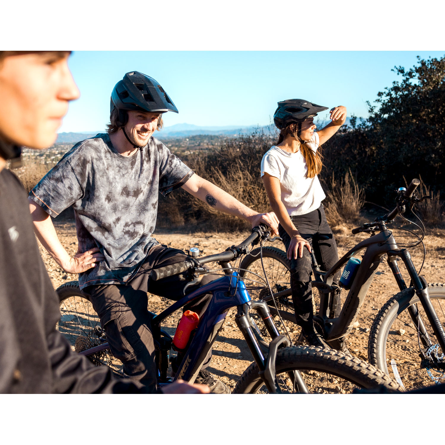 Trois cyclistes casqués sur des vélos sur un sentier de terre, avec un arrière-plan panoramique.