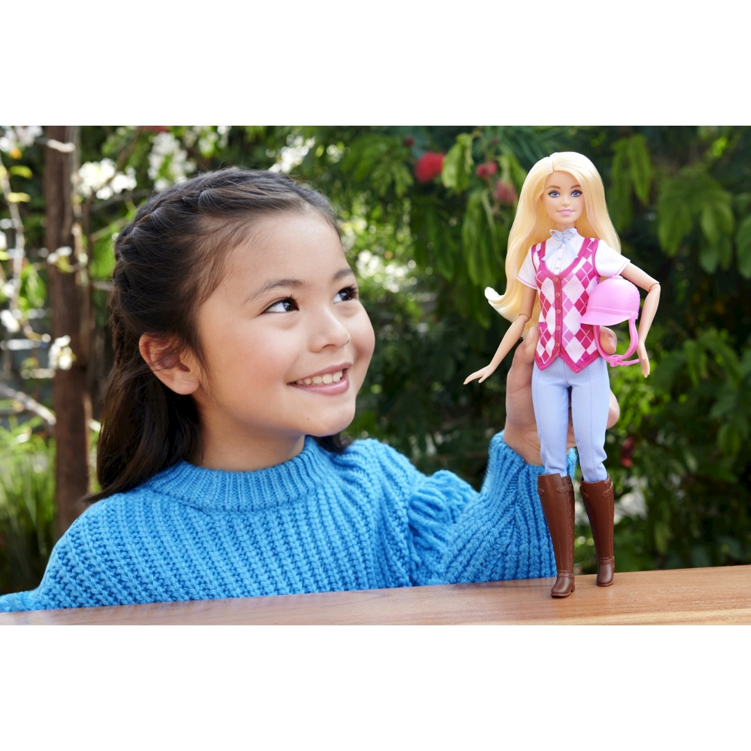 La ragazza tiene in mano una bambola Barbie con abiti da equitazione. Lo sfondo mostra alberi e cielo.