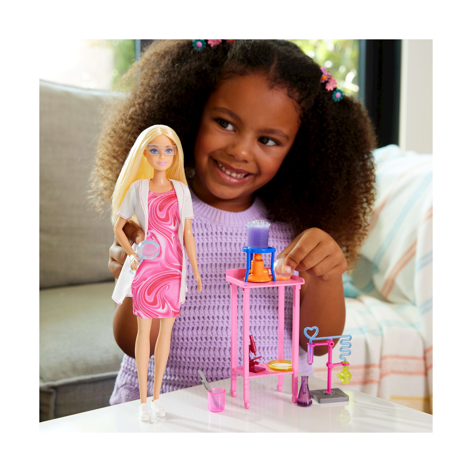 Una bambina sorride mentre tiene una bambola Barbie e un set da laboratorio scientifico con provette, becher e una fiala viola.