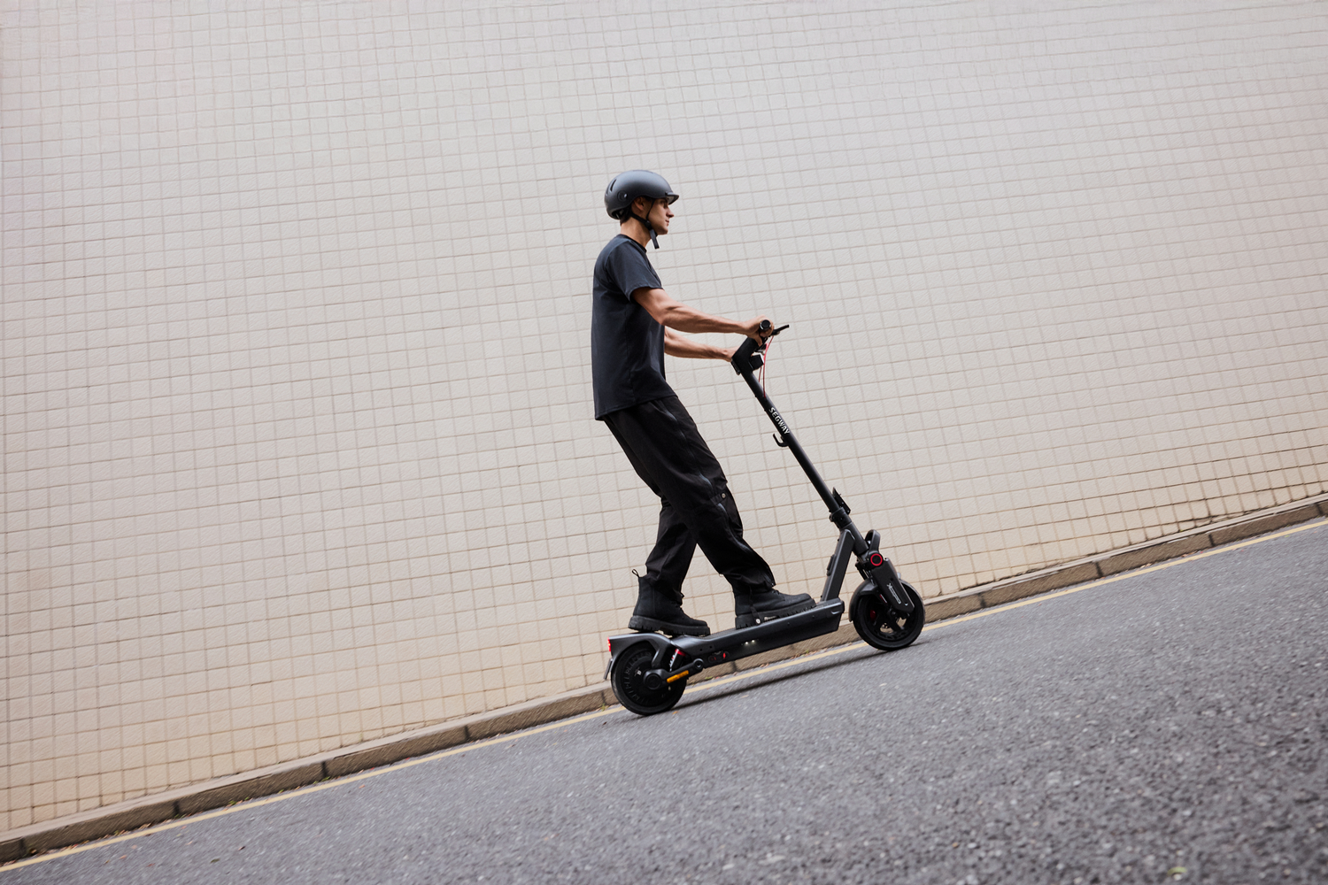 Una persona en un scooter eléctrico en una carretera junto a una pared. La persona lleva casco.