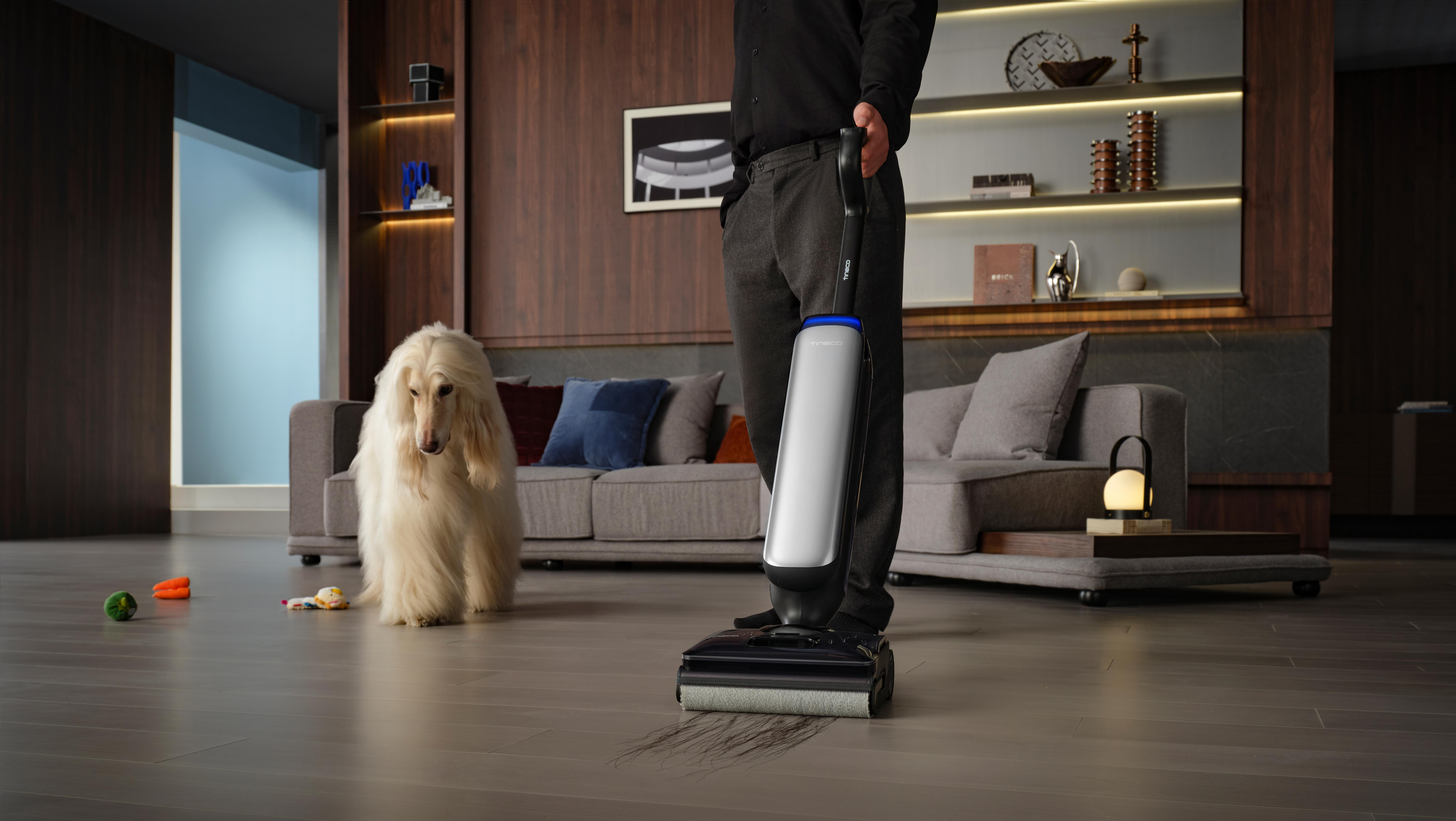 Ein Mann benutzt einen Staubsauger, ein Hund sitzt daneben. Wohnzimmer mit dunklem Holz und Sofa.