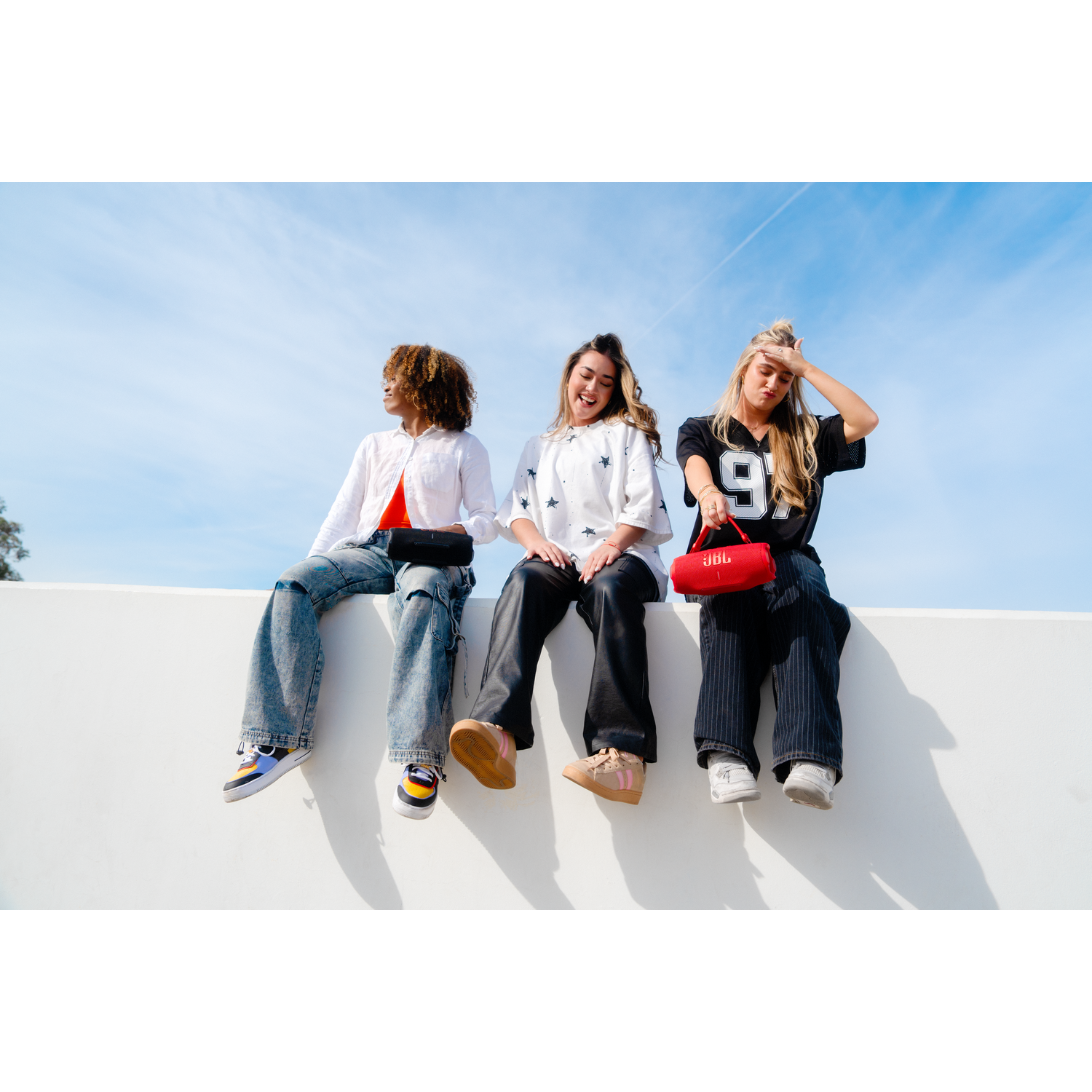 Three women sitting on a white wall, holding a red speaker, under a blue sky.