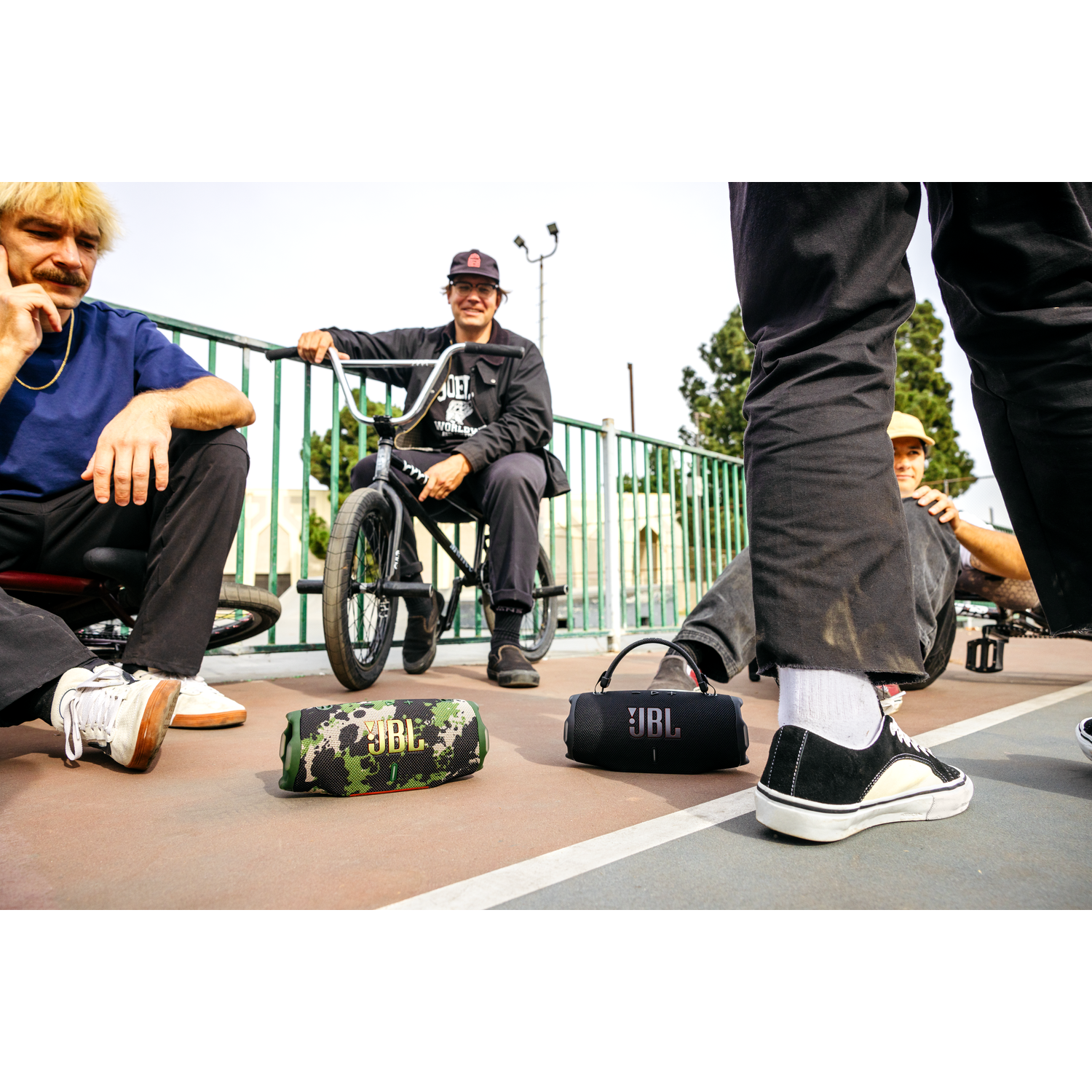 Group of young men with two JBL speakers, one camouflaged, one black, on a basketball court, bikes nearby.