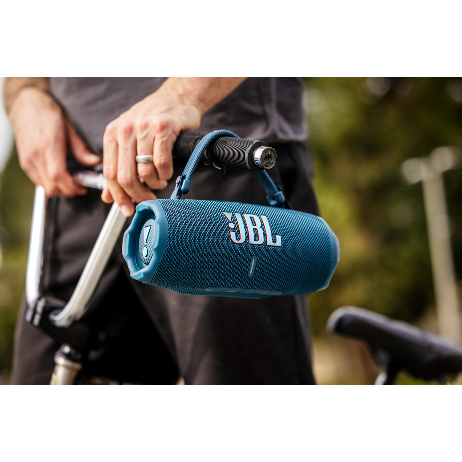 Blauer JBL-Lautsprecher wird von einer Person auf einem Fahrrad gehalten. Der Lautsprecher hat einen blauen Griff.