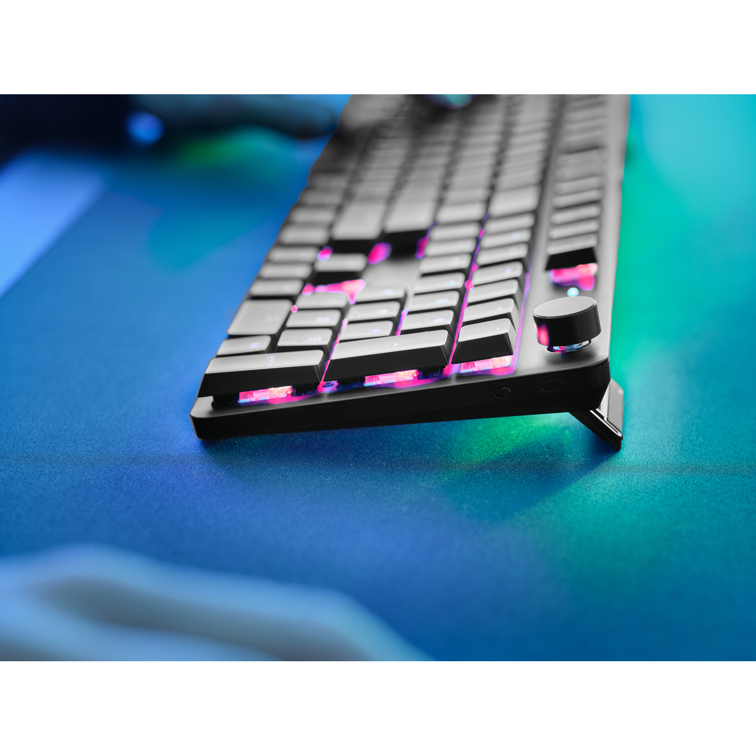 A close-up of a backlit black keyboard with pink and white keys. There is a volume knob.