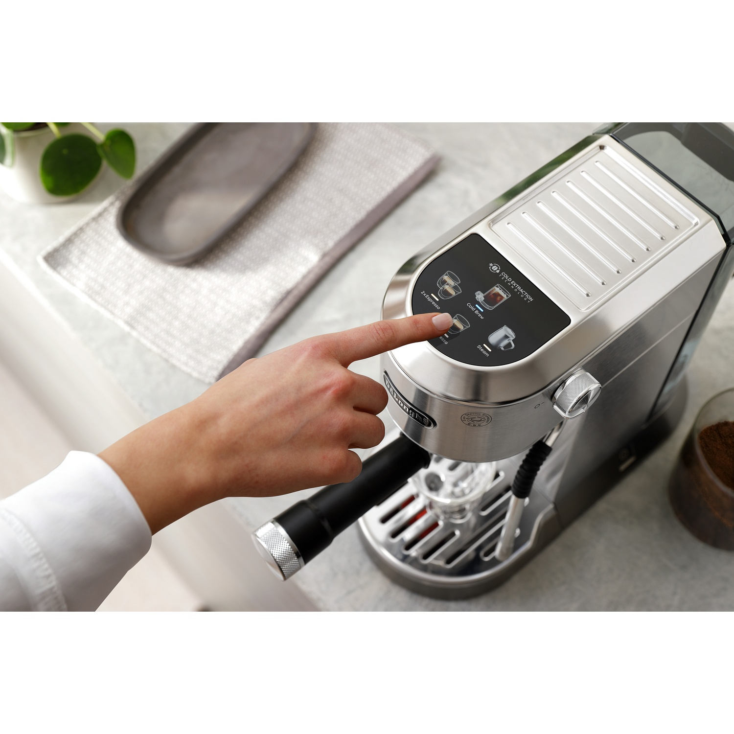 A person's hand presses a button on a silver espresso machine. Coffee cup and beans are nearby.