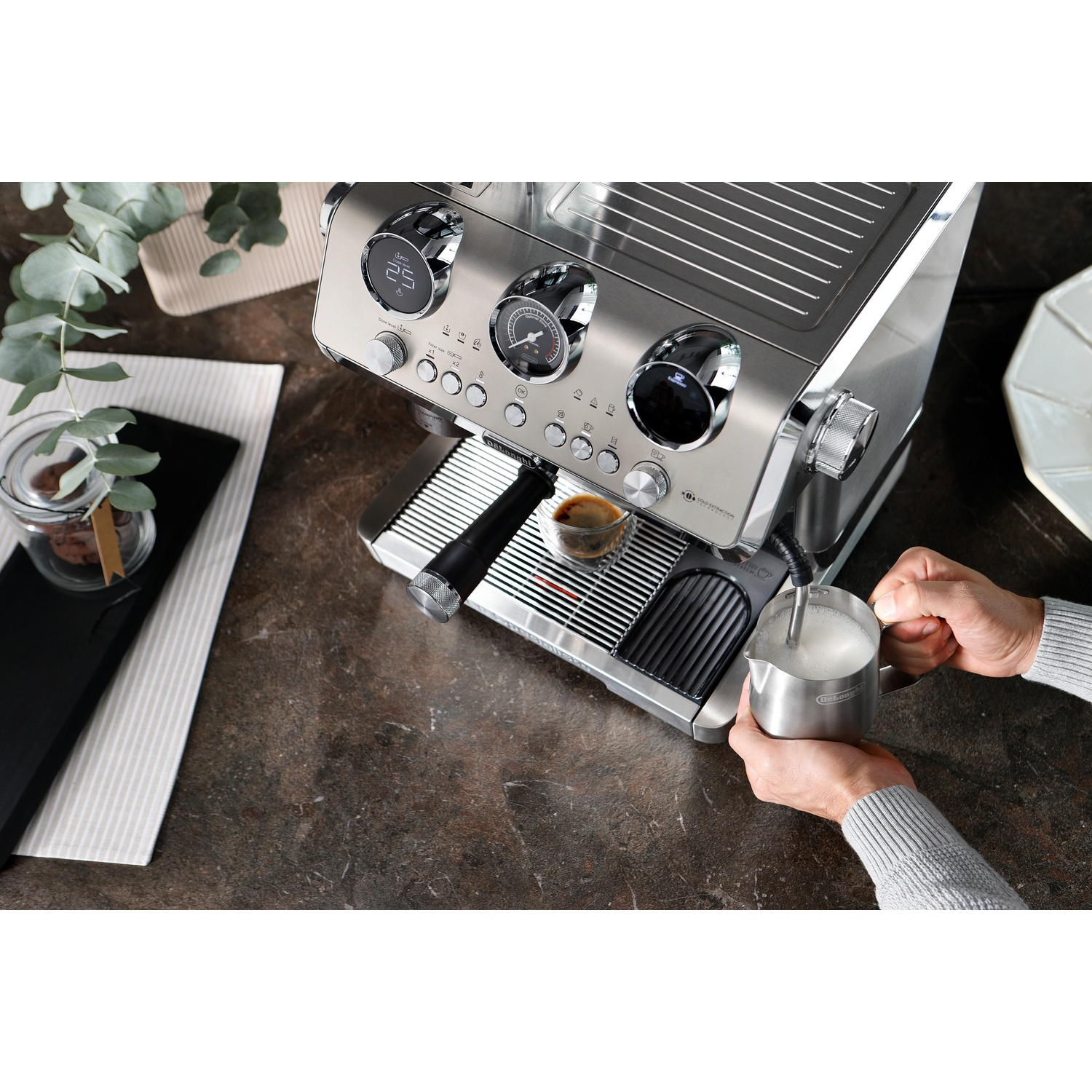 Espresso machine with a cup, a milk frothing jug, and a person's hands.
