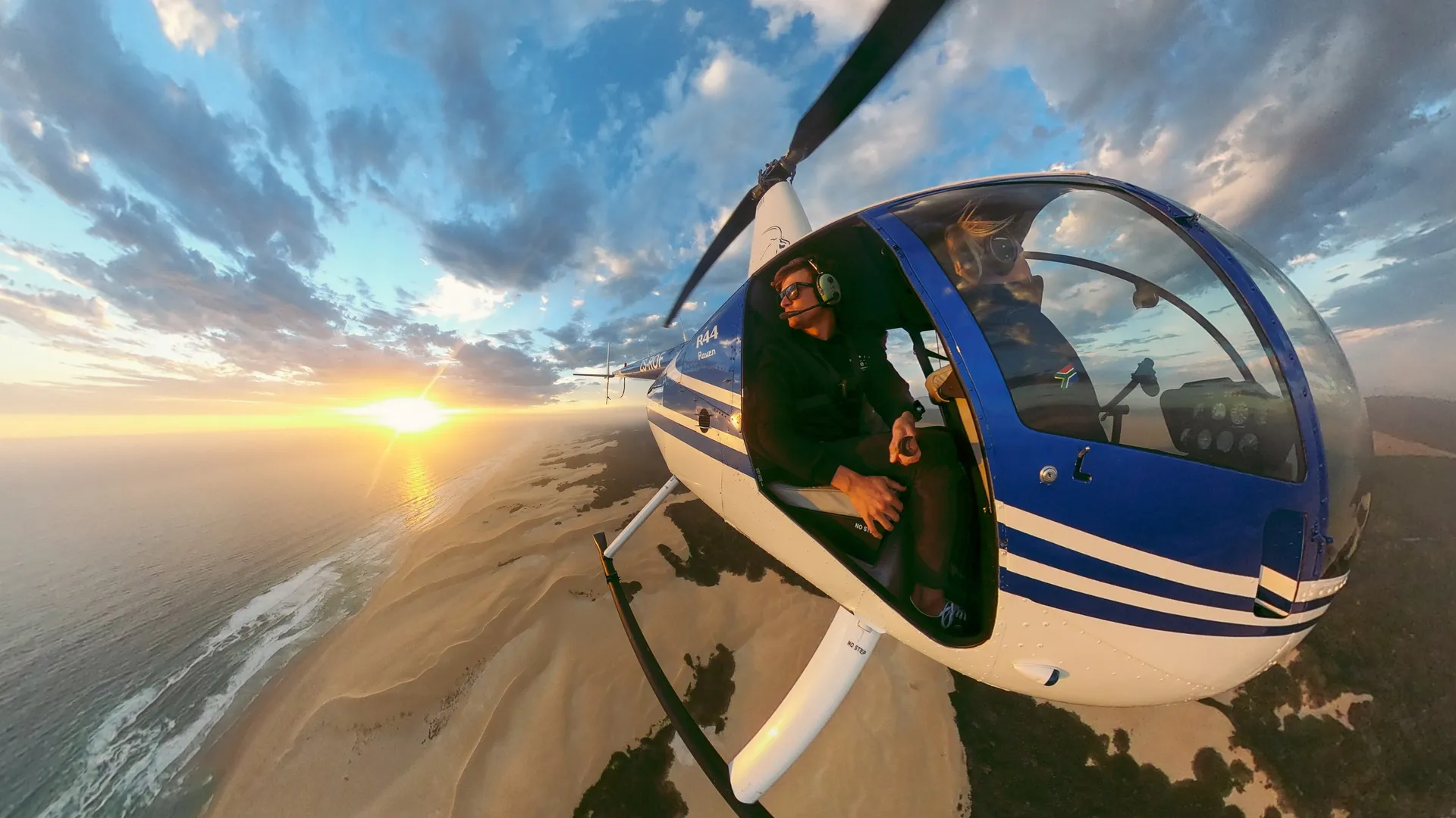 Un elicottero in volo sull'oceano al tramonto, con un pilota visibile.