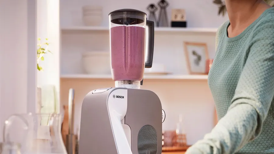 Un mixeur avec un smoothie rose est sur un comptoir de cuisine. Une femme se tient à proximité.