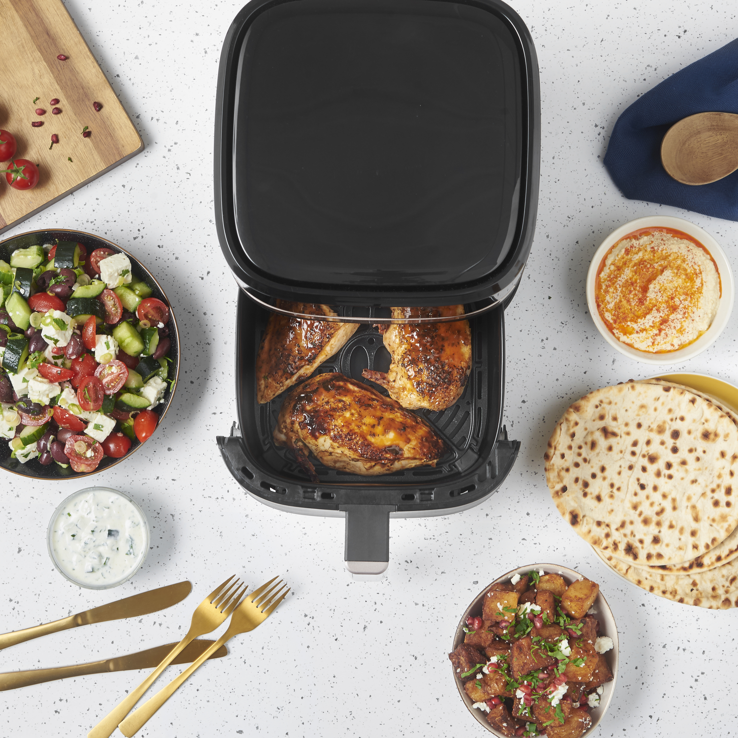 An overhead shot shows a black air fryer with cooked chicken. Various dishes surround it: salad, hummus, pita, potatoes.