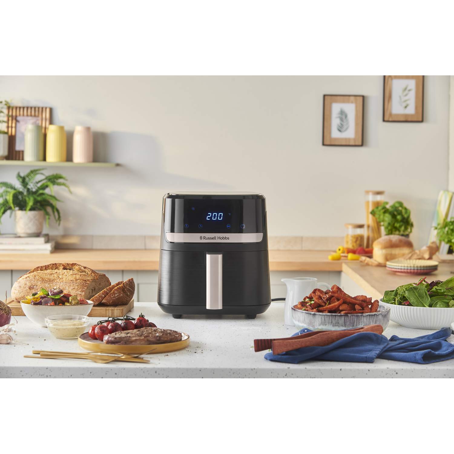 An air fryer on a countertop with food and utensils. Bread, salad, tomatoes, and other ingredients are also visible.