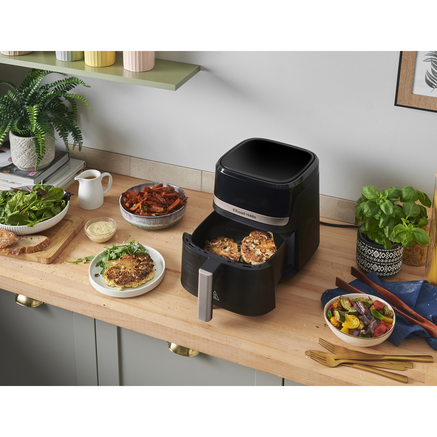 An air fryer with vegetables, and cooked food. Bowls of food, plants, and utensils are on a wooden countertop.