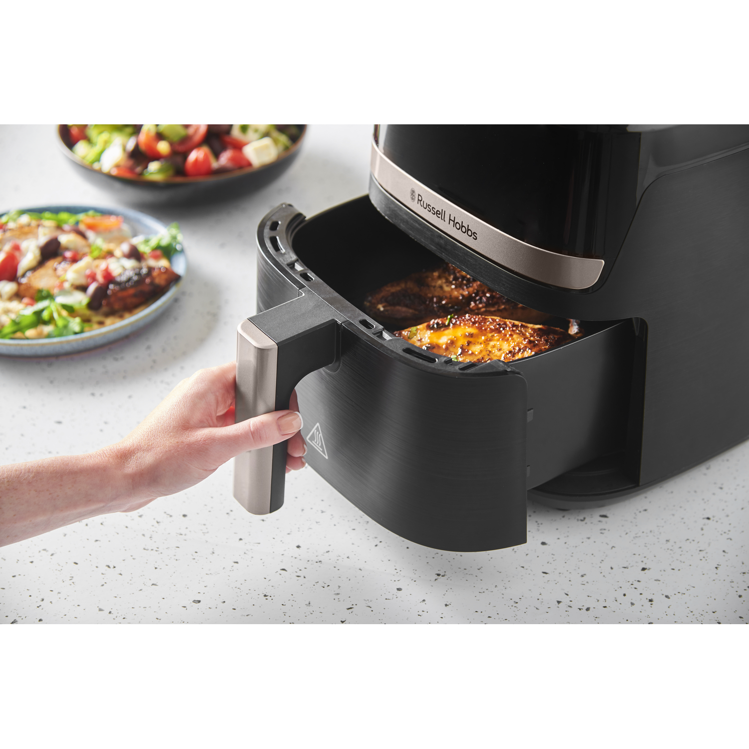 A black air fryer with food inside. A hand pulls out the basket. Salads are in the background. Russell Hobbs logo.