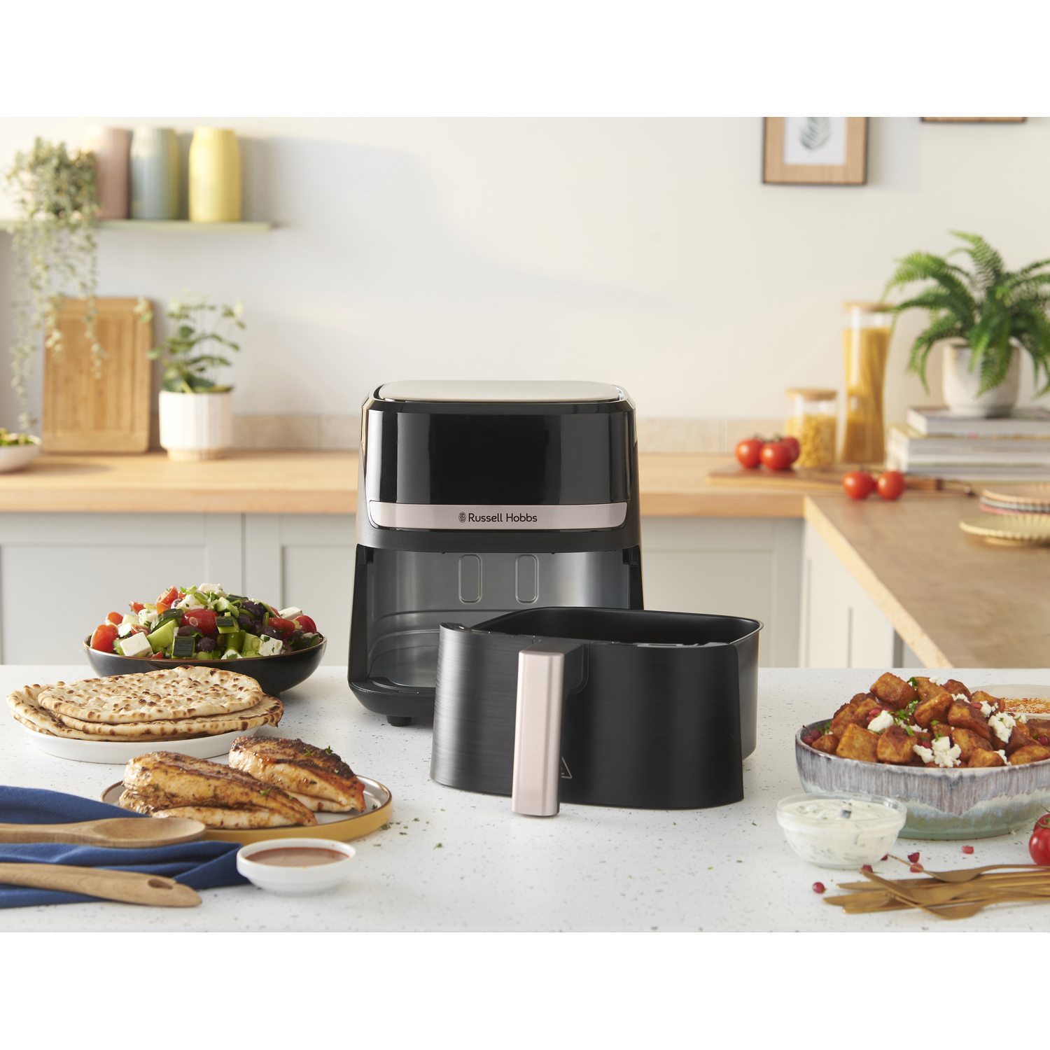 An air fryer with food: salad, pita, chicken, and potatoes. Background has a kitchen counter, plants, and jars.